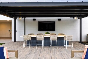 An outdoor patio with a black pergola, a white brick wall, and a flat-screen TV mounted above a bar area with four wicker chairs and a central table.