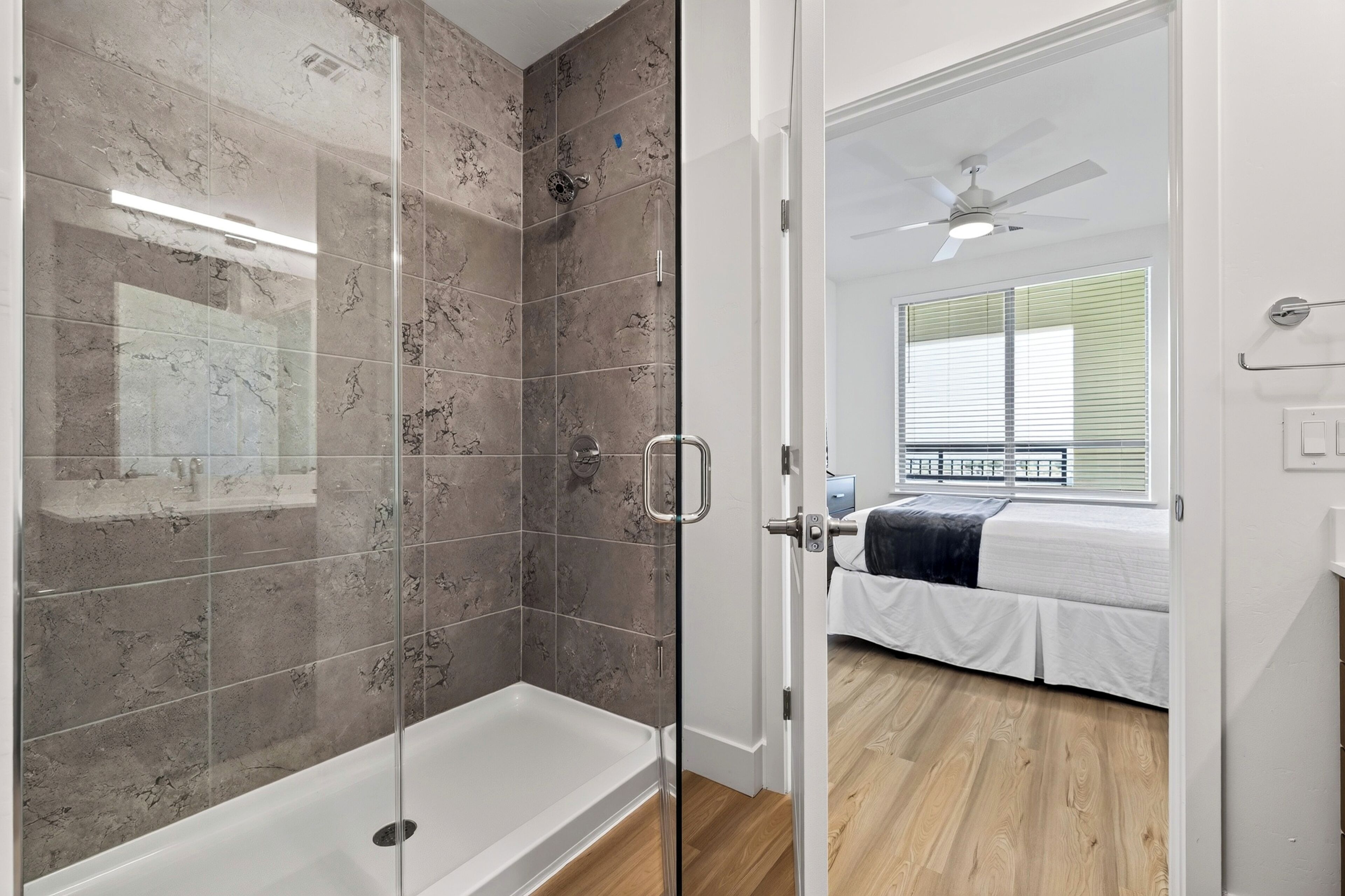 A modern bathroom with a glass shower enclosure, a white bathtub, and a wooden floor leading to a bedroom with a bed and a ceiling fan.