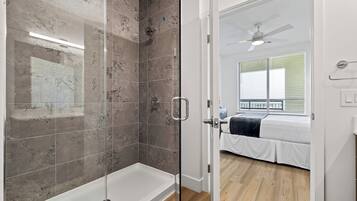 A modern bathroom with a glass shower enclosure, a white bathtub, and a wooden floor leading to a bedroom with a bed and a ceiling fan.