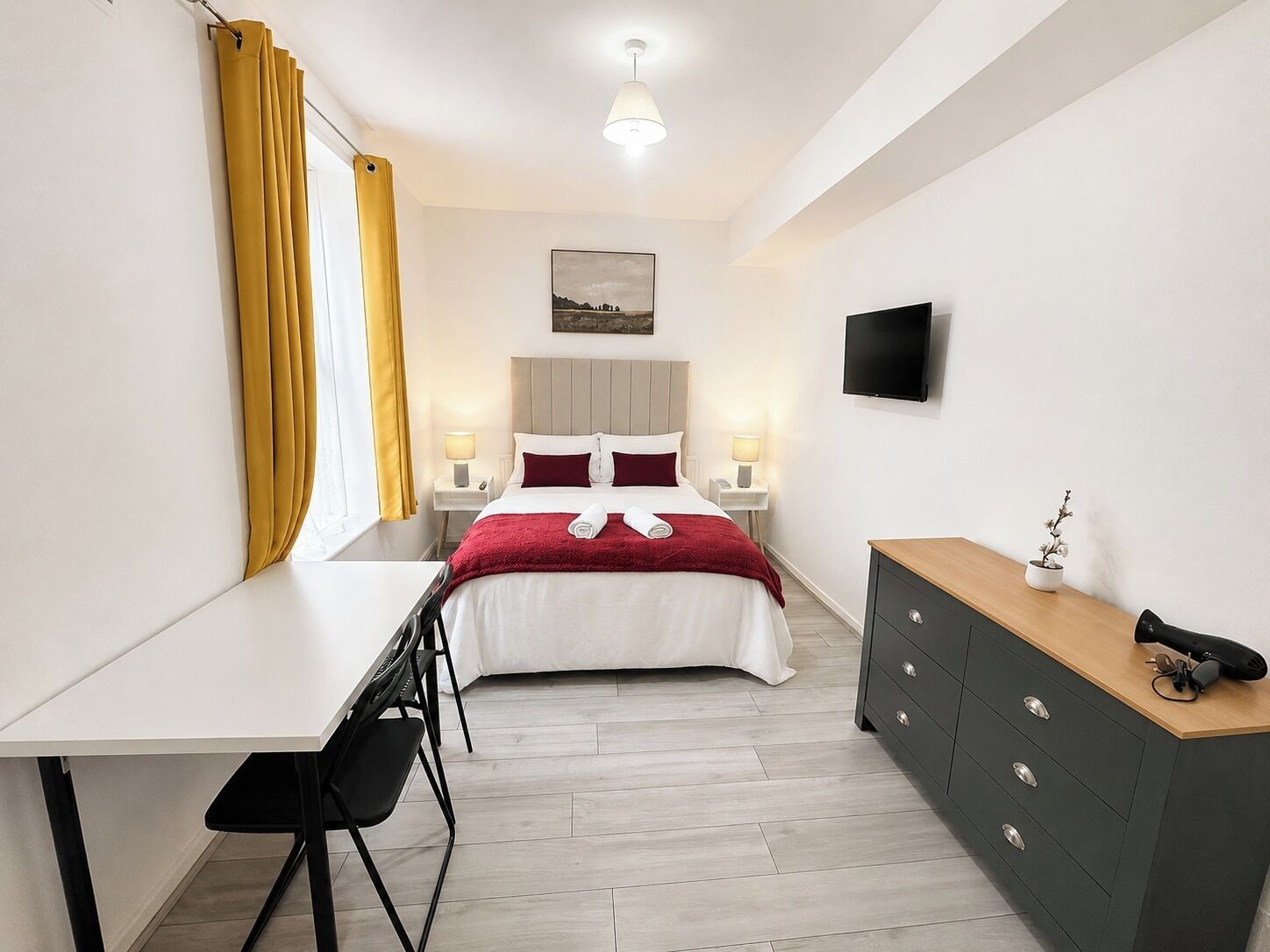 Ein Hotelzimmer mit Bett, Nachttischen, Schreibtisch, Kommode, Fernseher und einem Fenster mit Vorhängen.