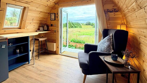 A cozy cabin interior with a wooden ceiling, a dining area with a table and chairs, a sofa, and a view of a green landscape through a large window.
