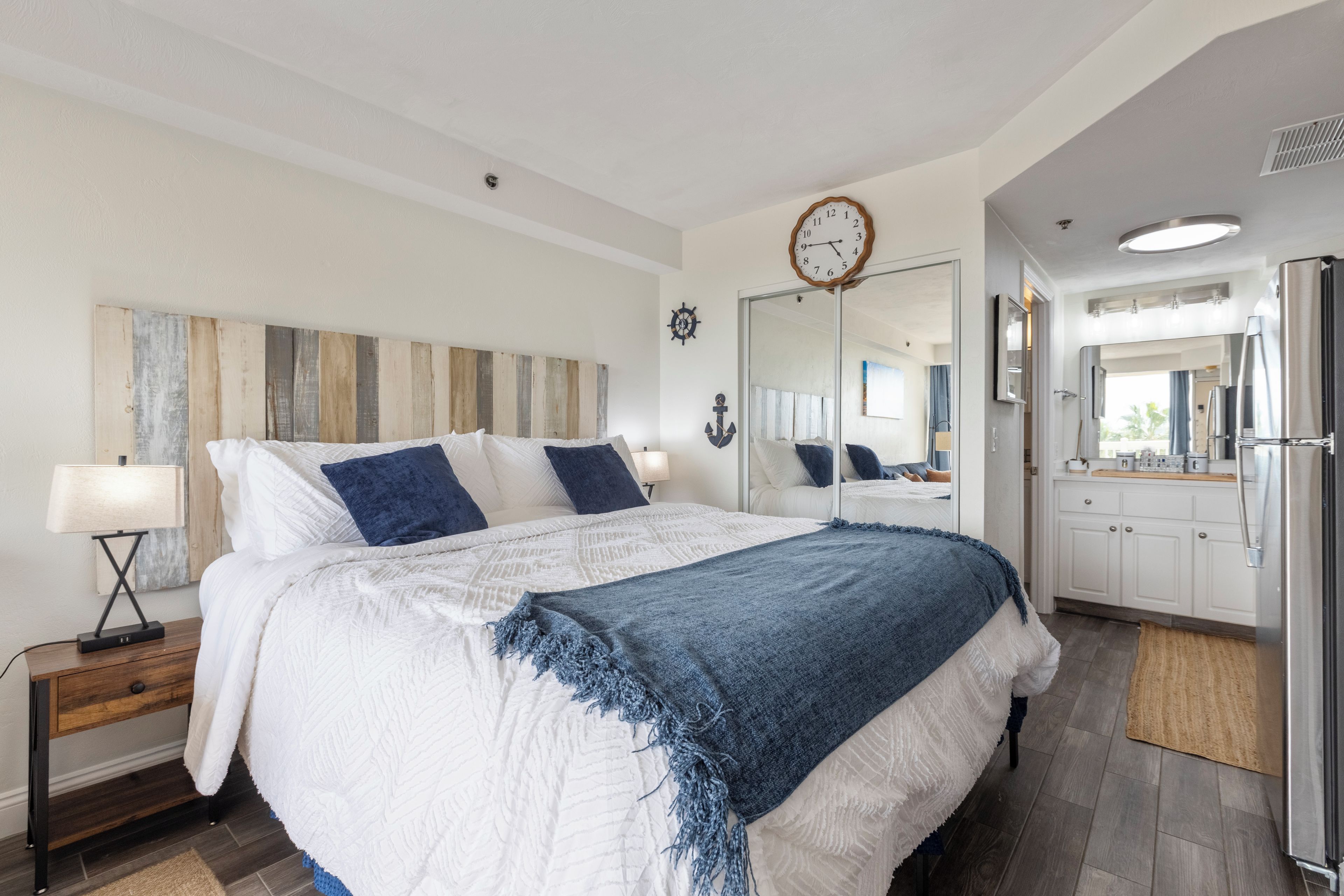 A modern bedroom with a large bed, bedside lamps, a clock, and a view into the adjacent bathroom.
