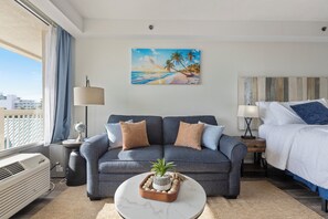 Ein Hotelzimmer mit grauem Sofa, einem kleinen runden Tisch mit Pflanze, einem Bild mit Strandmotiv und einem Bett mit weißer Bettwäsche.