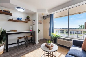 A modern apartment with a kitchen area, a dining table with two stools, a sofa, a small round table, and a large window with a view of the street. - New! Life’s a Beach! 1 BR King Suite Resort (Daytona Beach)