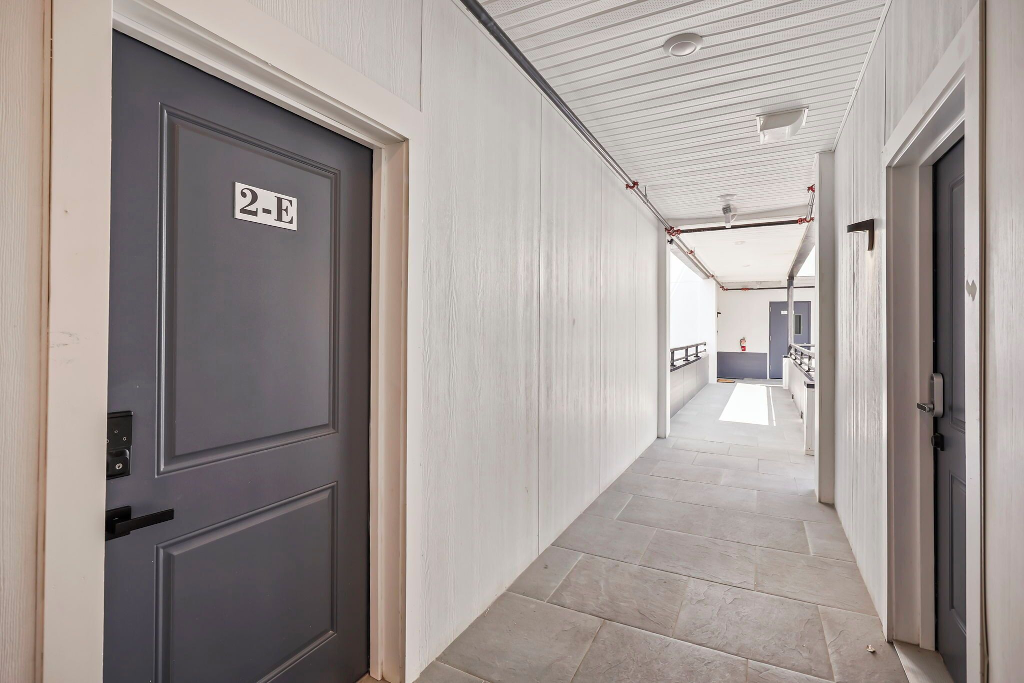 A hallway with a grey door labeled '2-E' and a white ceiling with a grid pattern.
