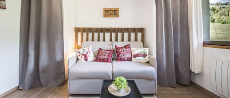 Ein modernes Wohnzimmer mit einer Couch, dekorativen Kissen, einem kleinen Tisch mit einer Pflanze und einem Fenster mit Vorhängen.