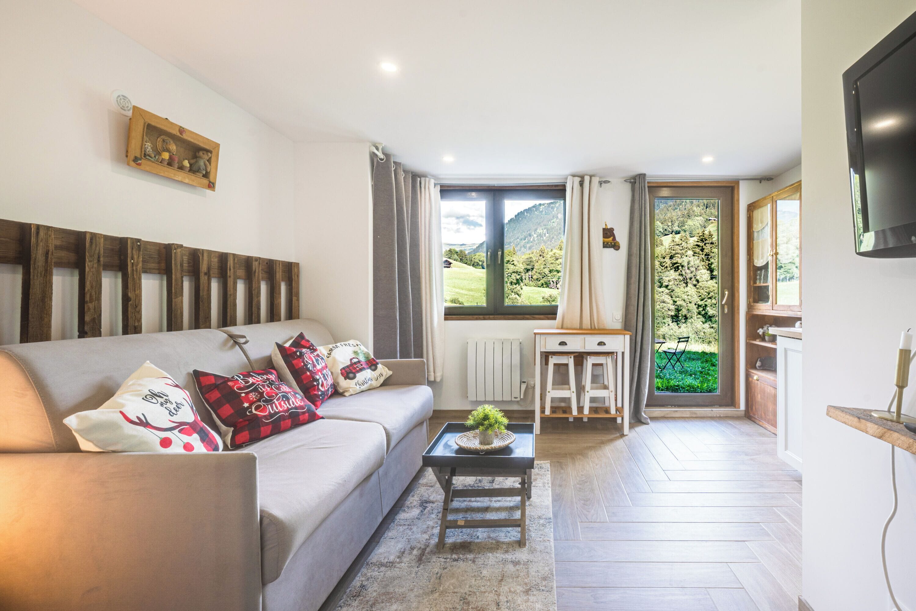 Una sala de estar con un sofá, una mesa de centro con una planta, un televisor y vistas a un paisaje verde a través de una ventana.