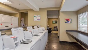A laundry facility with multiple washing machines, a seating area with a TV, and a window with blinds. - Riverview Condo by Downtown - Pool, Free Covered Parking & Patio (Nashville)