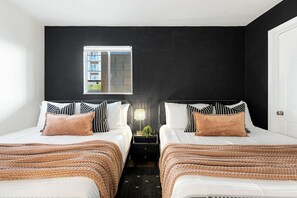 A hotel room with two beds, a nightstand, a lamp, and a window with a view of buildings.