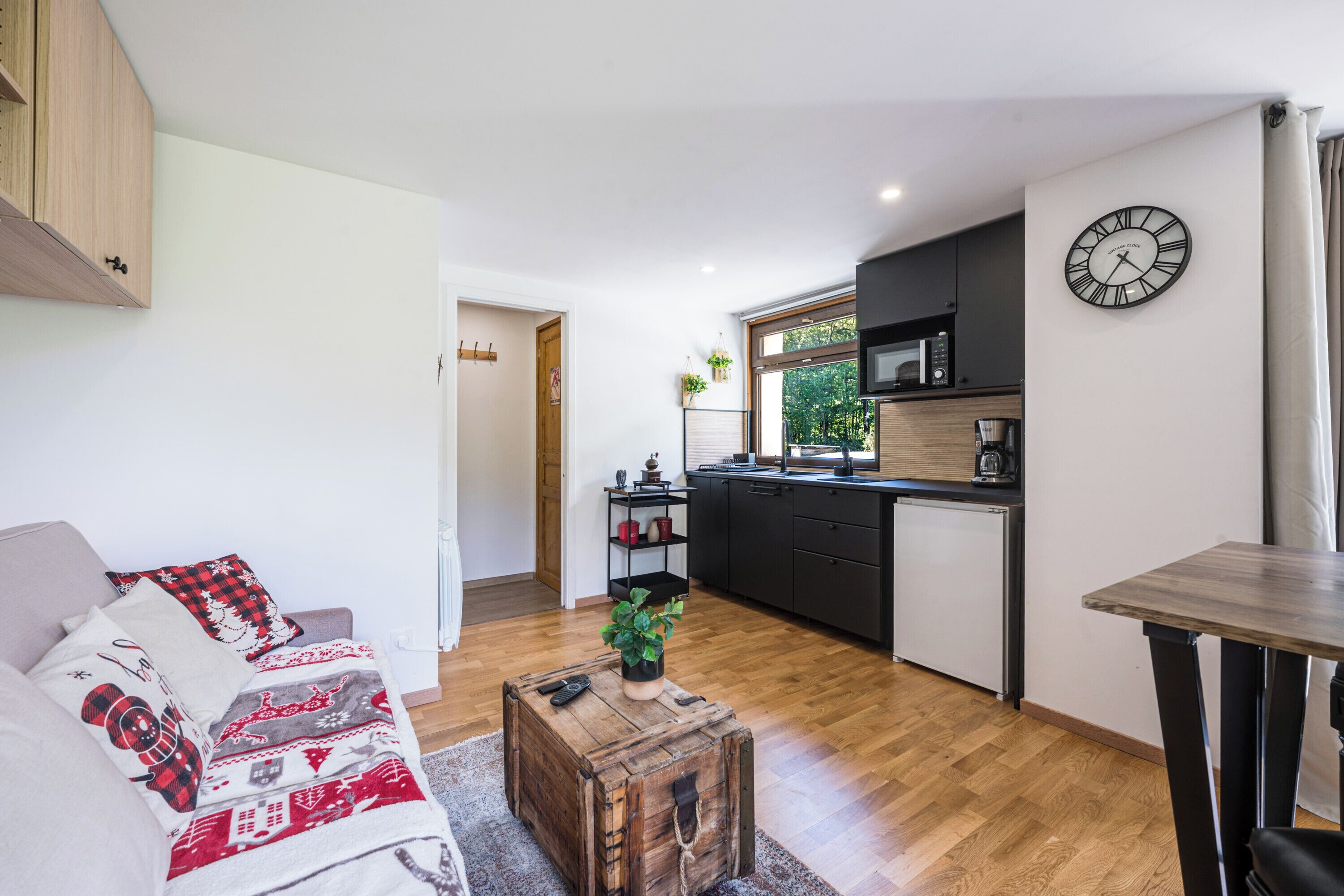 A compact living space with a kitchenette, a sofa with patterned cushions, a wooden coffee table, and a clock on the wall.
