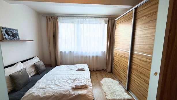 A bedroom with a bed, a bedside table, a window with curtains, a wooden wardrobe, and a small rug. - Modern 1,5 - room apartment, Kadnárova 91 - Rača (Bratislava)
