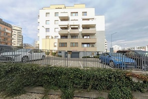 Ein mehrstöckiges Wohngebäude mit geschlossenem Eingangsbereich, geparkten Autos und einer Hecke im Vordergrund.