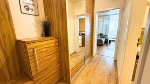 A hallway with a wooden cabinet, a mirror, and a door leading to a living area with a sofa and a table. - Modern 1,5 - room apartment, Kadnárova 91 - Rača (Bratislava)
