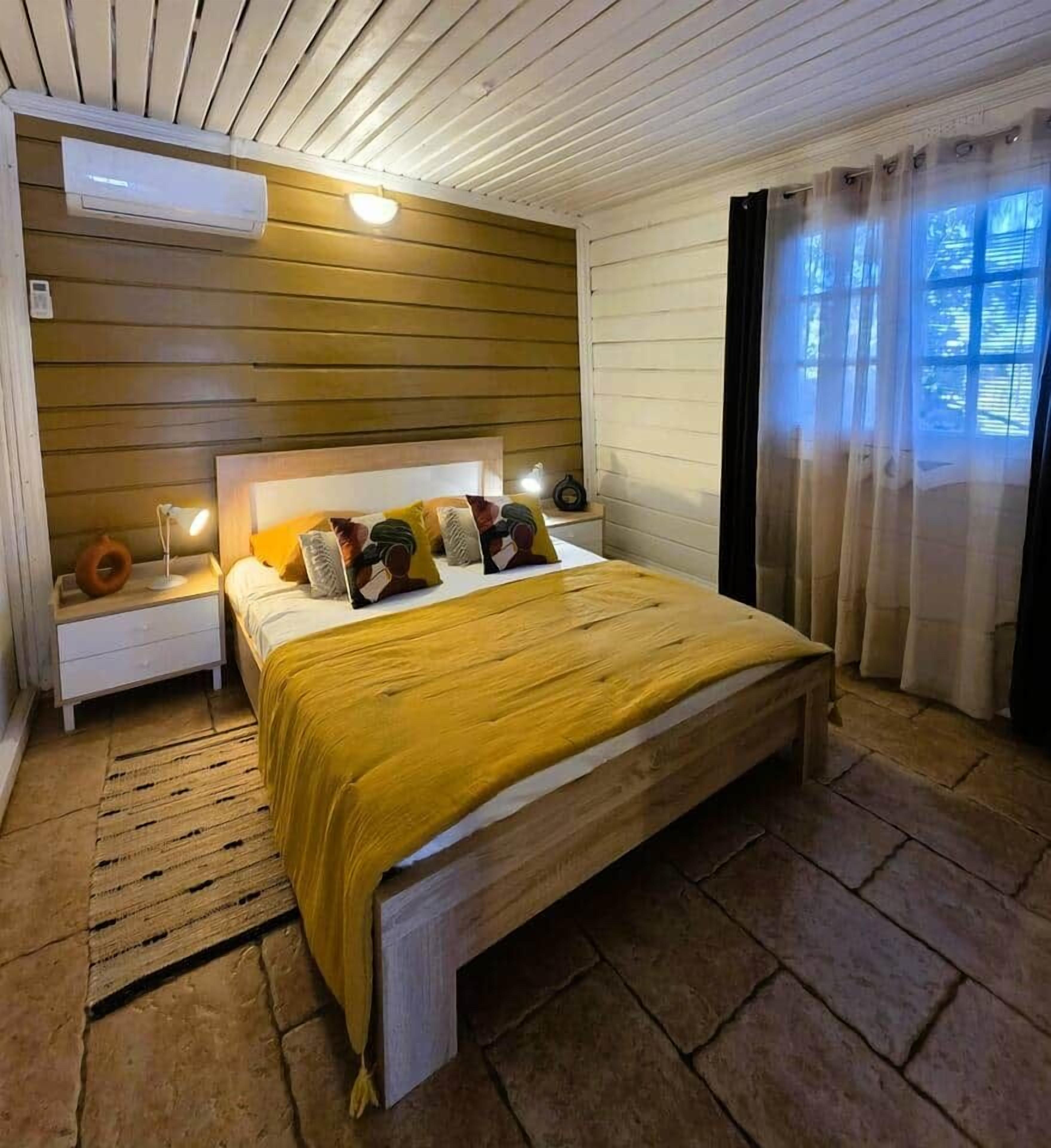 A bedroom with a wooden headboard, a bed with a yellow bedspread, a nightstand, and a window with curtains.