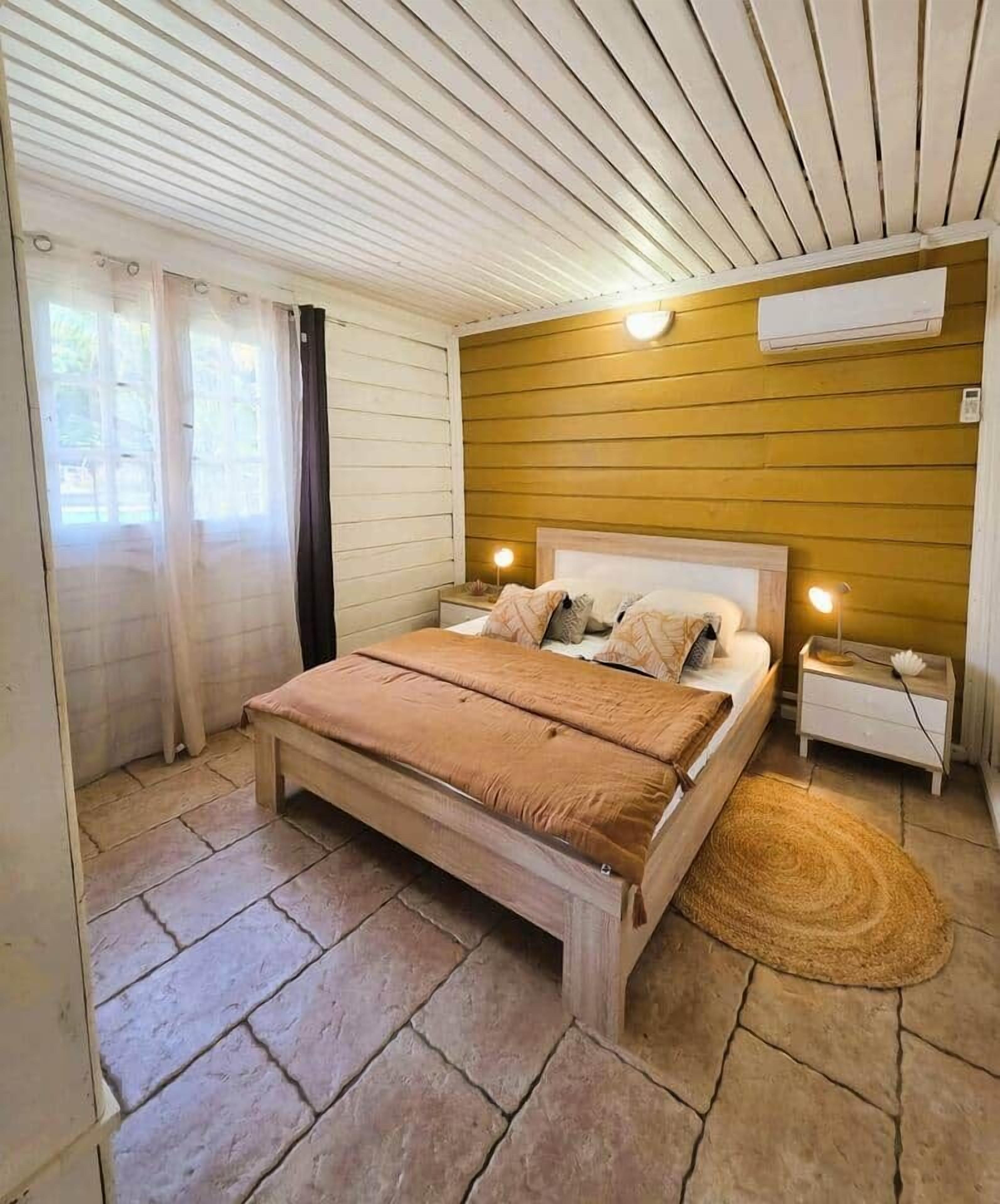 A bedroom with a bed, bedside tables, an air conditioning unit, and a window with curtains.