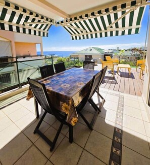 Ein Balkon mit Tisch und Stühlen, Blick auf das Meer und ein überdachter Bereich mit gestreiftem Markisenstoff.