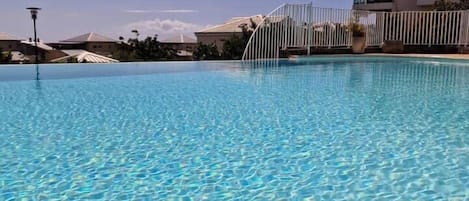 Uma piscina translúcida com grade e edifícios ao fundo.