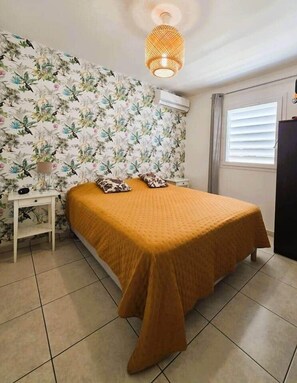 Ein Schlafzimmer mit einem Bett, einem Nachttisch, einer blumenmusterigen Wand, einem Fenster mit Jalousien und einer Pendelleuchte.