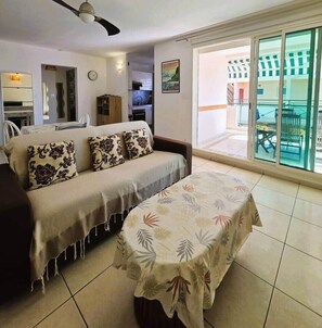 Sala de estar com sofá, mesa e vista para área externa através de uma porta de vidro.
