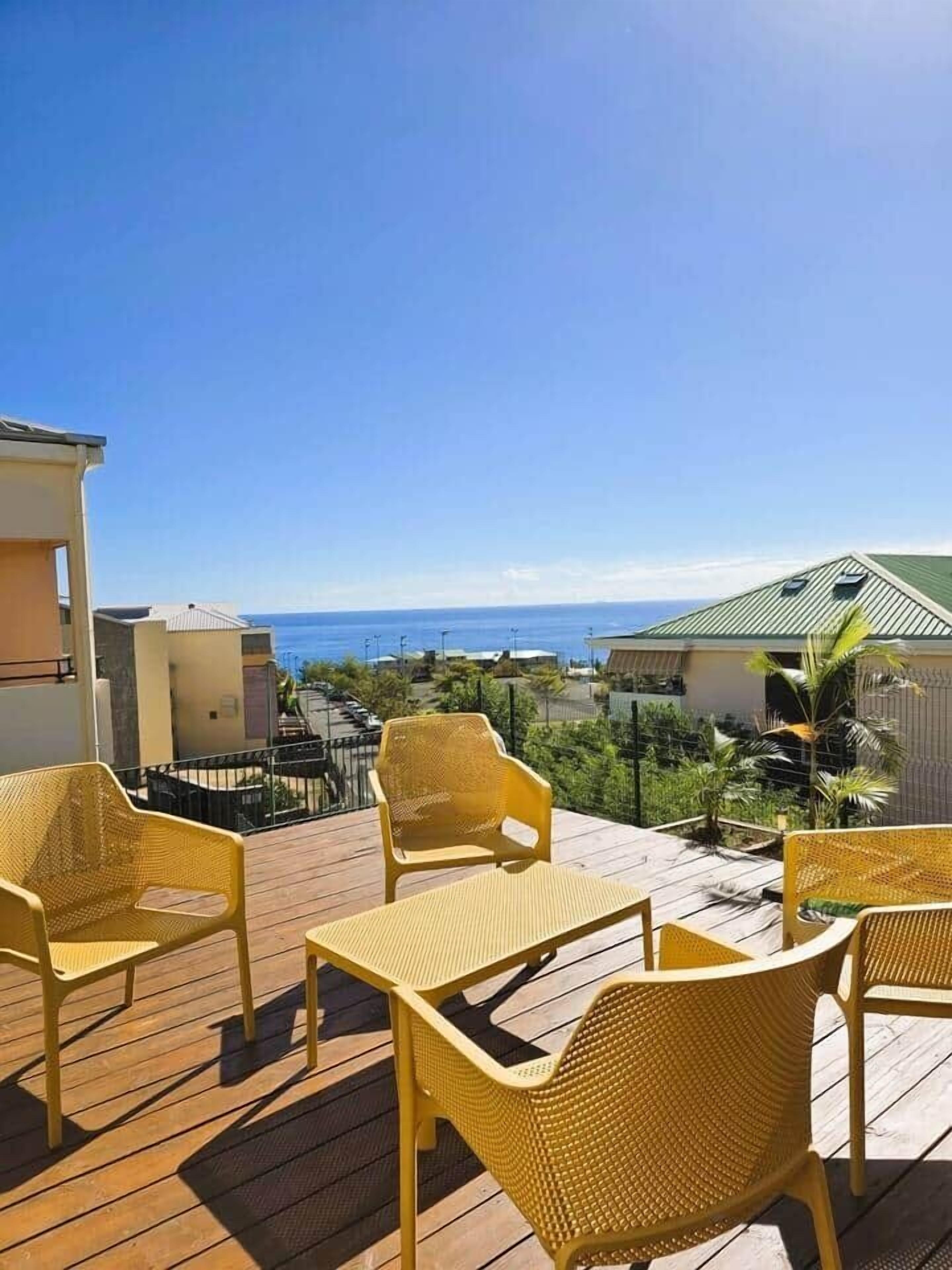 A wooden deck with yellow wicker chairs and a table, overlooking a residential area and the ocean.