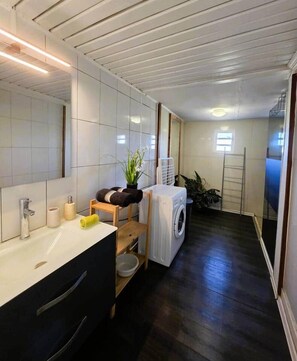 Une salle de bain moderne avec un grand miroir, des carreaux blancs, un lavabo, un lave-linge et une petite plante.