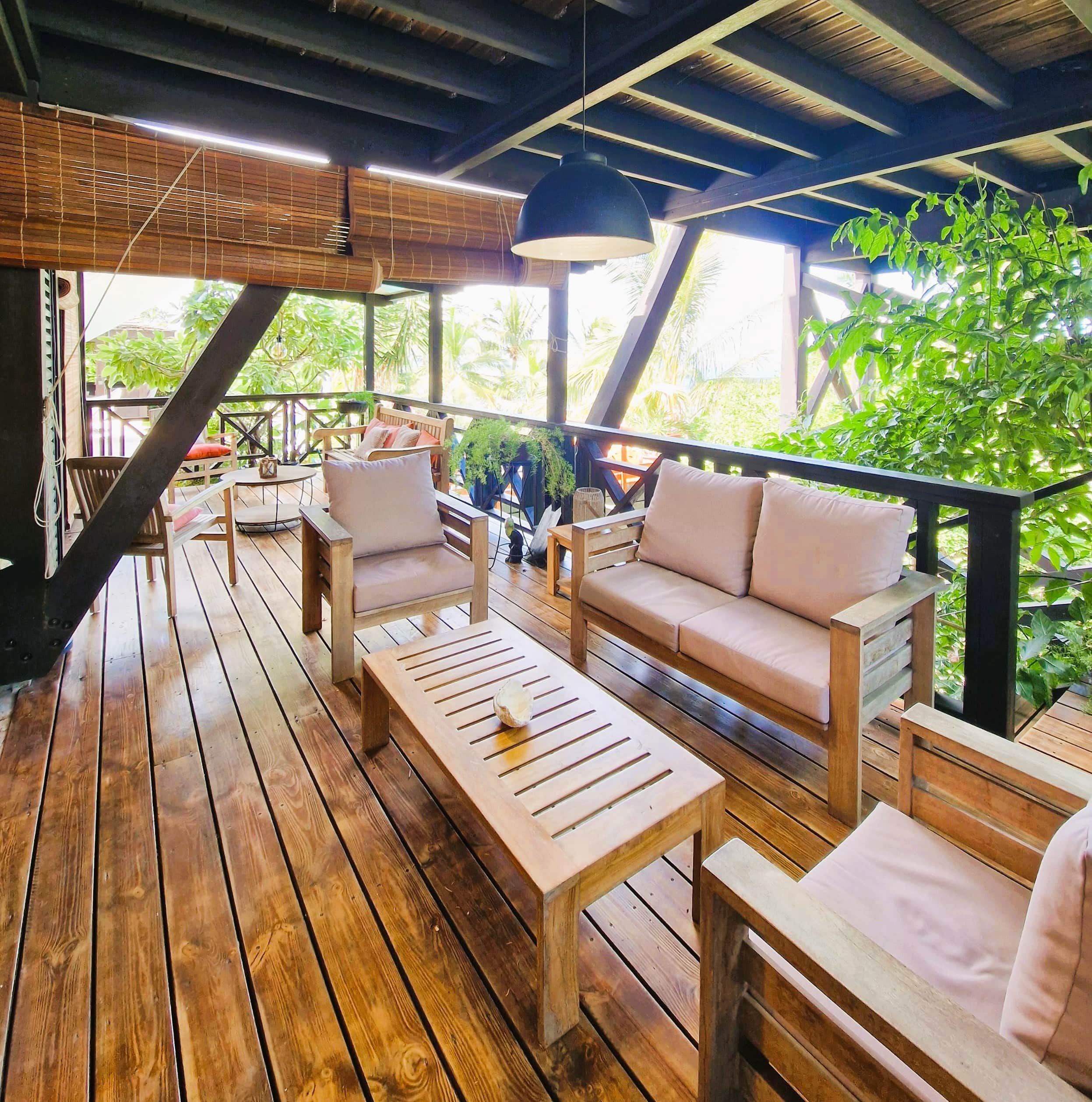 Una terraza exterior de madera con asientos, incluyendo un sofá y sillas, y una lámpara colgante.