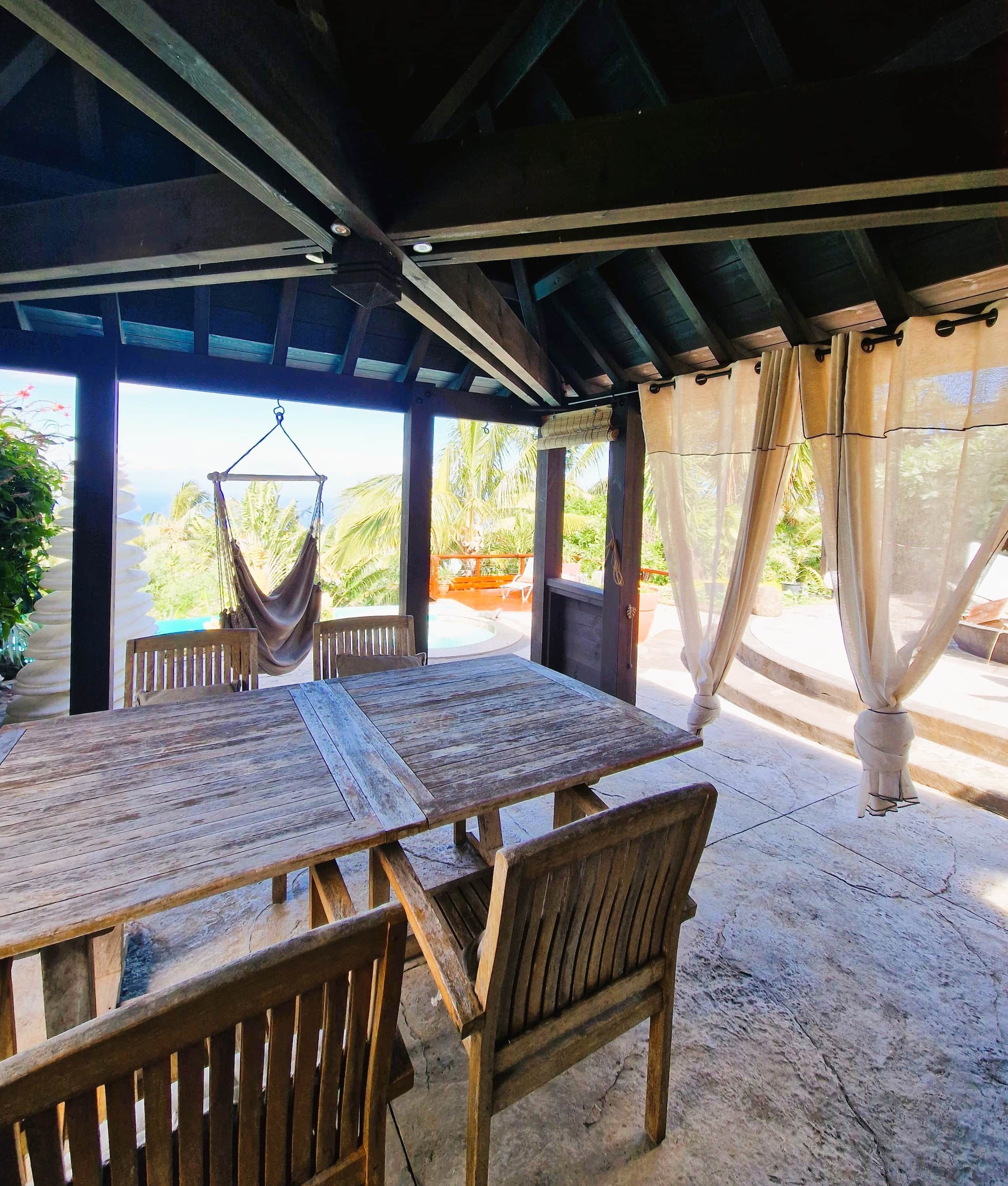 An outdoor covered dining area with wooden tables and chairs, a hammock, and a view of the ocean.