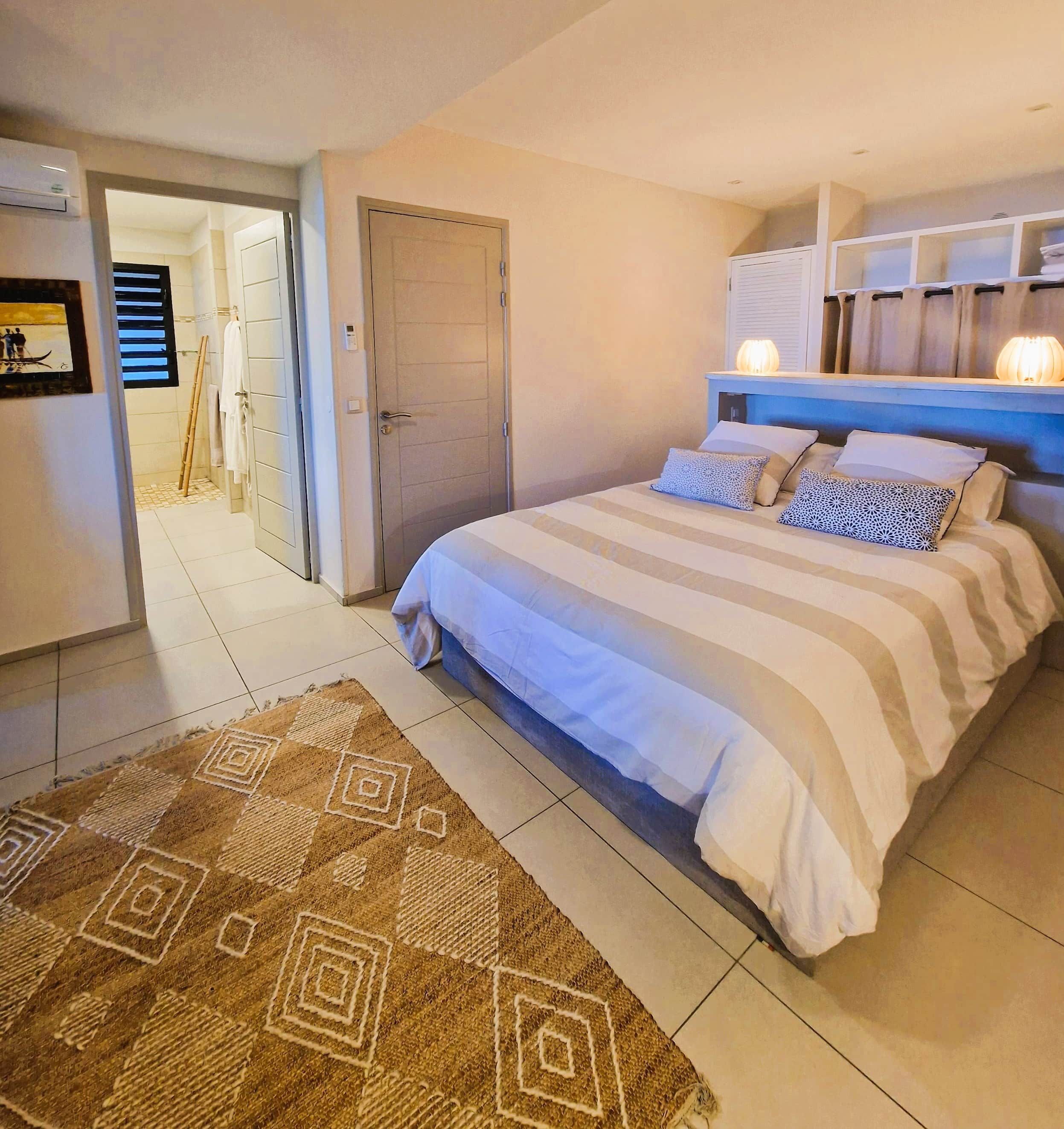 A bedroom with a bed, a nightstand, a wall-mounted shelf, a door, and a bathroom visible in the background.