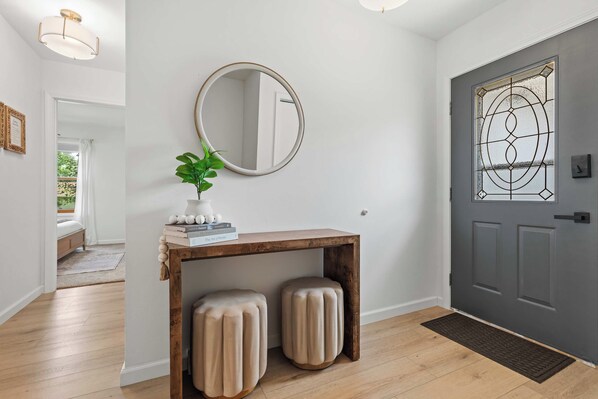 A hallway with a wooden console table, two cushioned stools, a round mirror, and a door with a decorative glass panel.