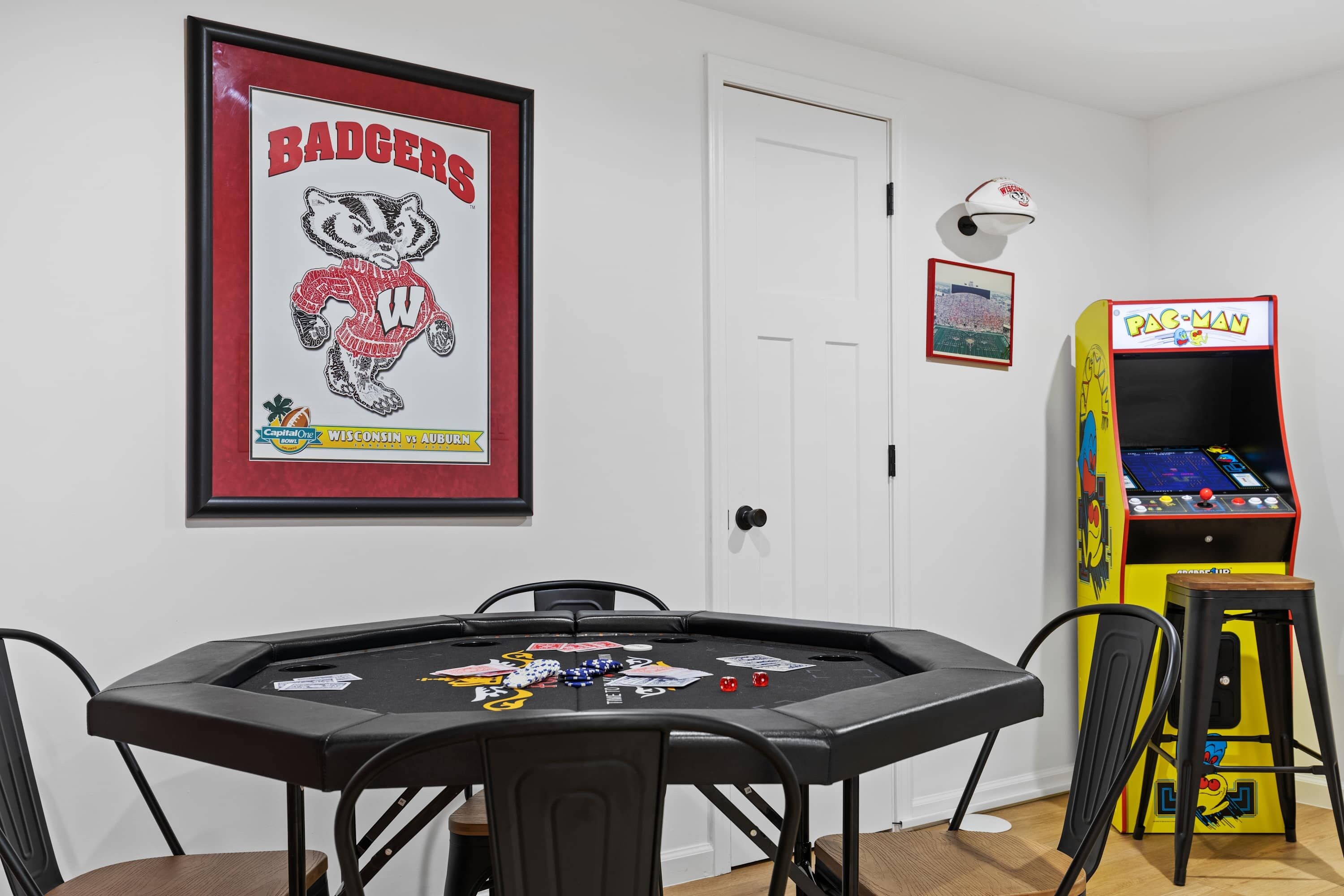 A room with a poker table, framed posters, a door, and a Pac-Man arcade machine.