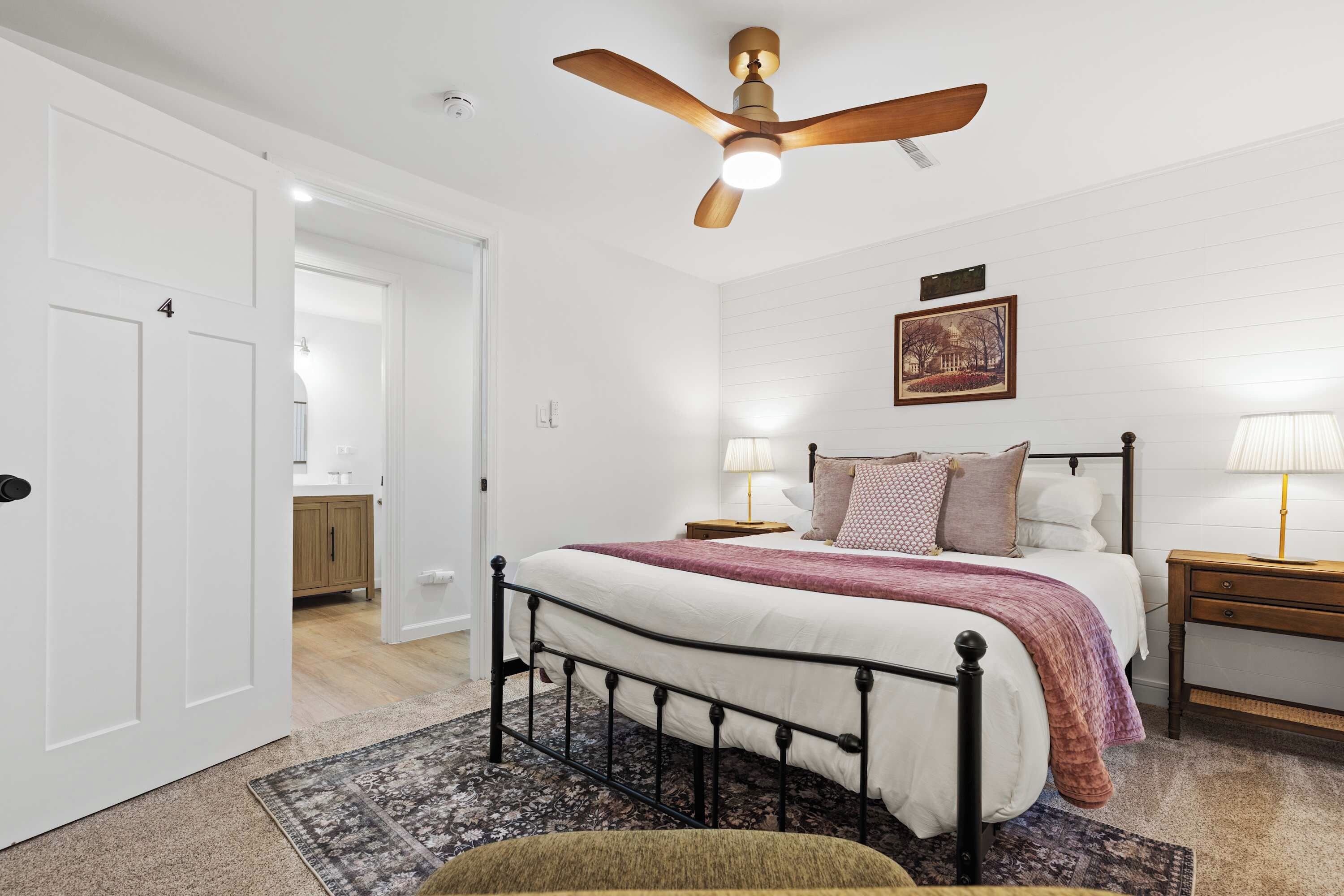 A bedroom with a bed, bedside tables, a ceiling fan, a framed picture on the wall, and a door leading to another room.