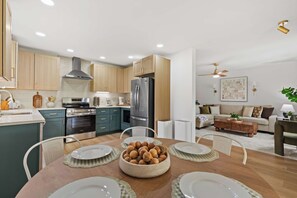 A modern kitchen with stainless steel appliances, a dining area with a wooden table, and a living room with a sofa and a ceiling fan.