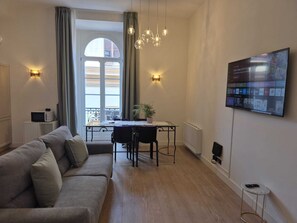 A modern living room with a sofa, dining table, and chairs. There is a TV mounted on the wall, a radiator, and a window with curtains. - NAPLES MARIANA LUXURY: SUPERIOR 2-BEDROOM APARTMENT IN THE CITY CENTER (Napoli)