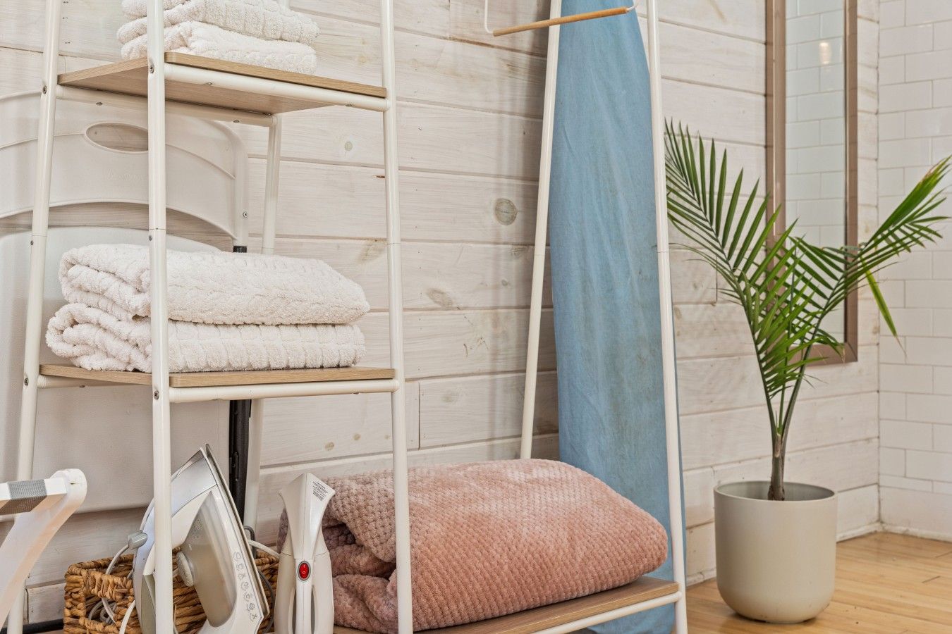 Een kamer met een witte metalen plank waarop opgevouwen handdoeken, een blauw gordijn, een pot met plant en een haardroger op de vloer staan.