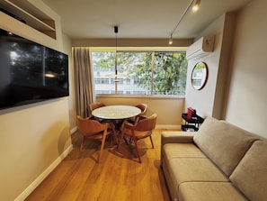 Ein modernes Wohnzimmer mit einem runden Esstisch, Ledersesseln, einem beigen Sofa, einem flachbildfernseher an der Wand und einem großen Fenster mit Blick auf Bäume und Gebäude.