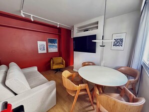 Ein modernes Wohnzimmer mit roter Akzentwand, einer Couch, einem gelben Sessel, einem runden Tisch mit Holzstühlen und einem an der Wand montierten Flachbildfernseher.