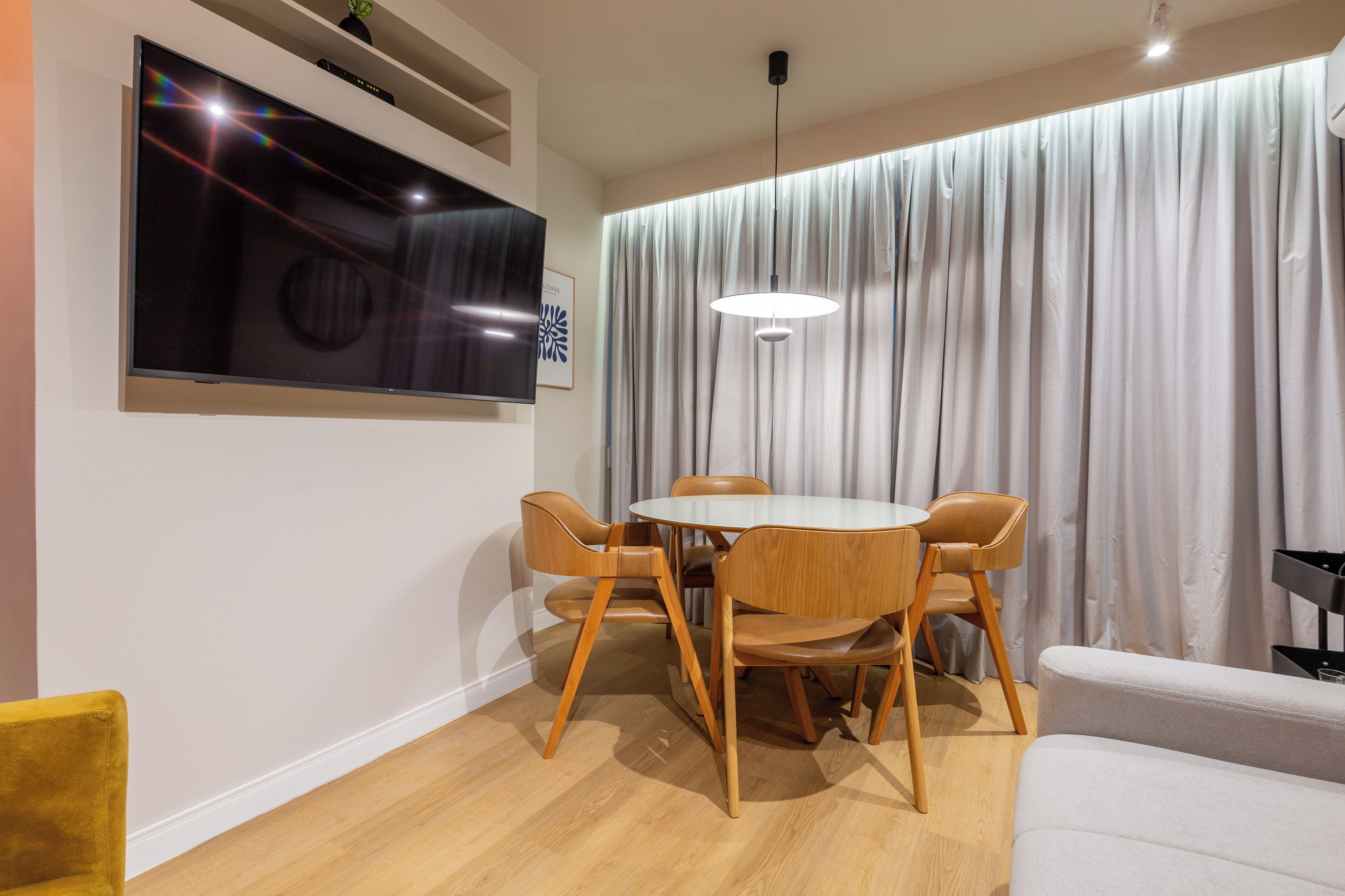 A modern dining area with a round table and four chairs, a flat-screen TV mounted on the wall, and a large window with curtains.