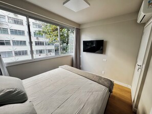Ein Hotelzimmer mit einem Bett, einem an der Wand befestigten Fernseher und einem Fenster mit Blick auf Gebäude und Bäume.