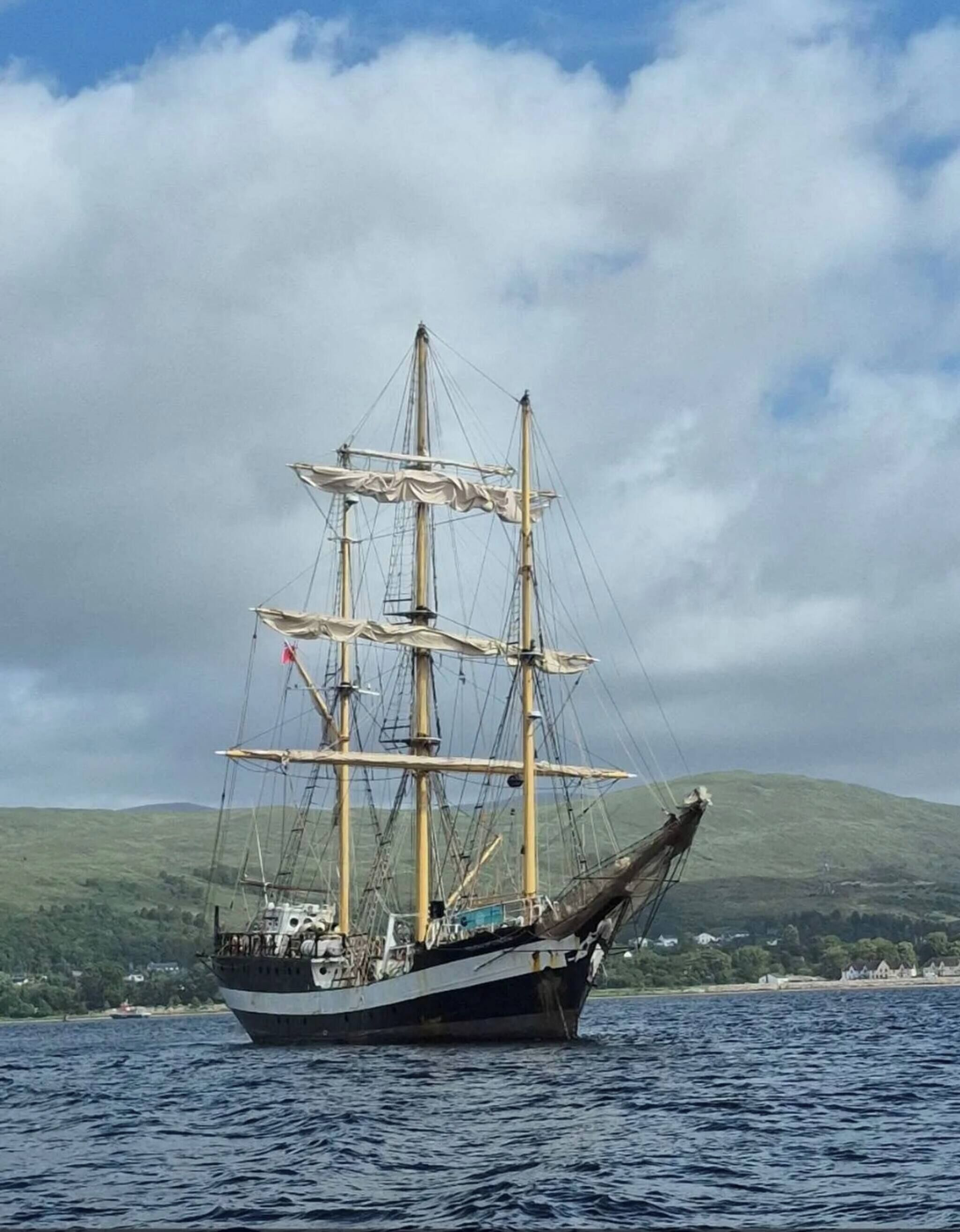 A tall ship with multiple masts and sails, anchored in a body of water with a hilly landscape in the background.