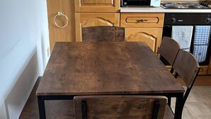 Uma área de refeições com mesa e cadeiras de madeira, uma cozinha com armários de madeira e micro-ondas, e piso revestido de azulejos.