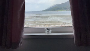 Finestra amb vista a una platja i muntanyes, cortines semiorades i un petit objecte decoratiu a la finestra.