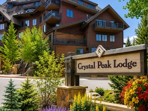 Un cartel de Crystal Peak Lodge con base de piedra, rodeado de follaje y un edificio de varios pisos al fondo.