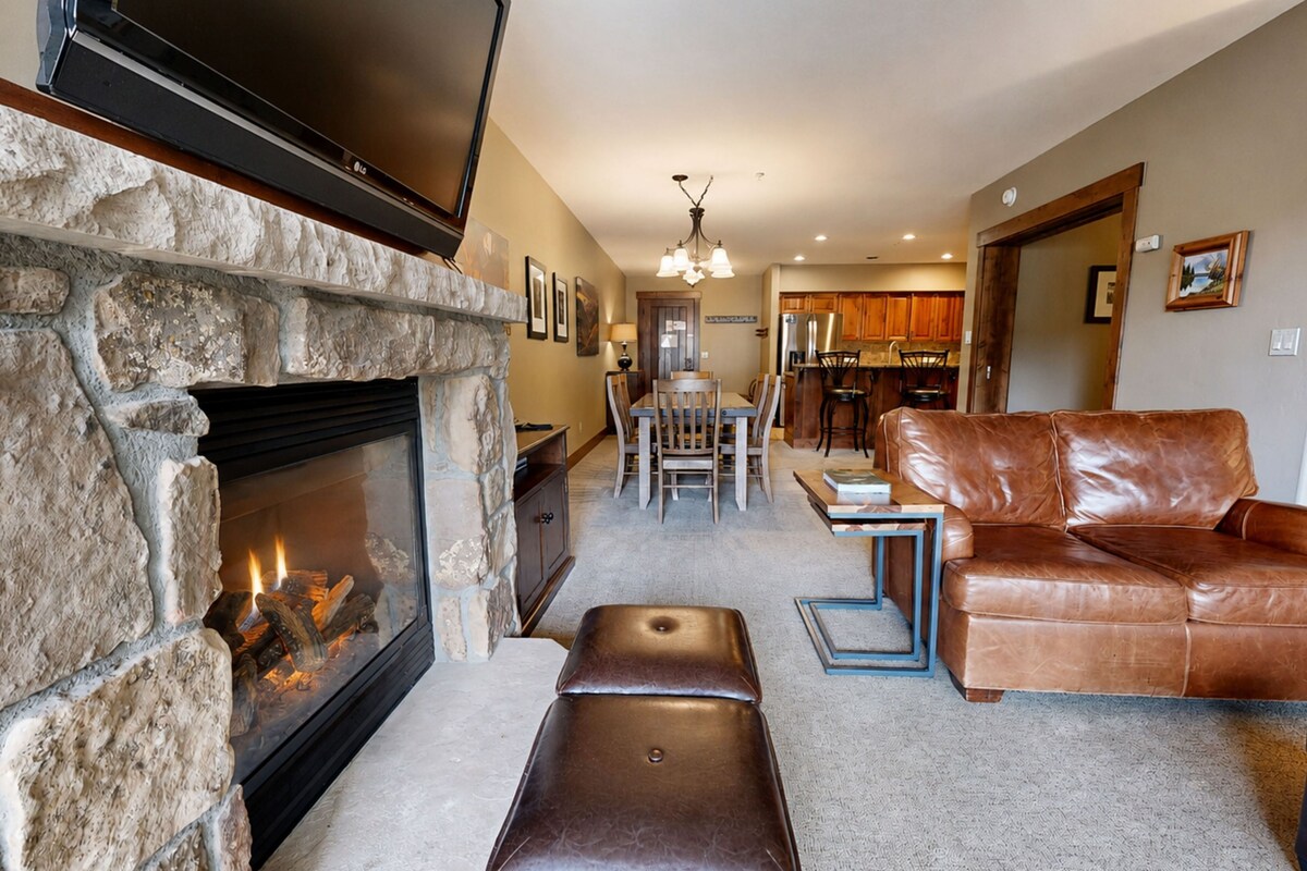 A living room with a stone fireplace, leather sofa, and a dining area with wooden chairs.