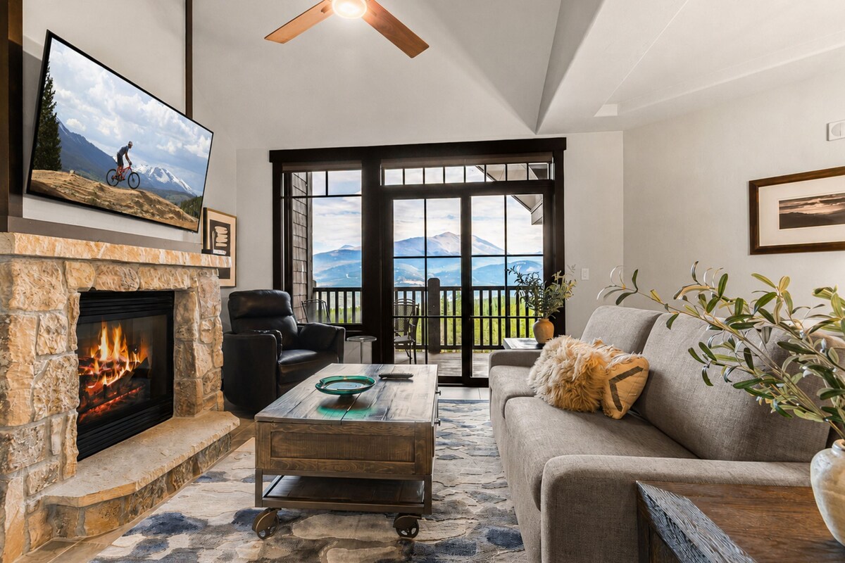 A living room with a stone fireplace, a television mounted above it, and a large window with a view of mountains.