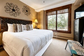 A bedroom with a large bed, a dresser, a TV, and a window with a view of trees. - Crystal Peak Lodge by CoralTree Residences (Breckenridge)