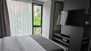 A hotel room with a bed, a television, a wardrobe, and a window with a view of buildings and trees.
