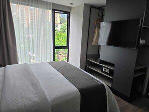 A hotel room with a bed, a television, a wardrobe, and a window with a view of buildings and trees. - BLOSS HOTEL (Medellín)