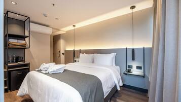 A modern hotel room with a neatly made bed, a minibar, a wall-mounted heater, and a bedside table with a cup and saucer.