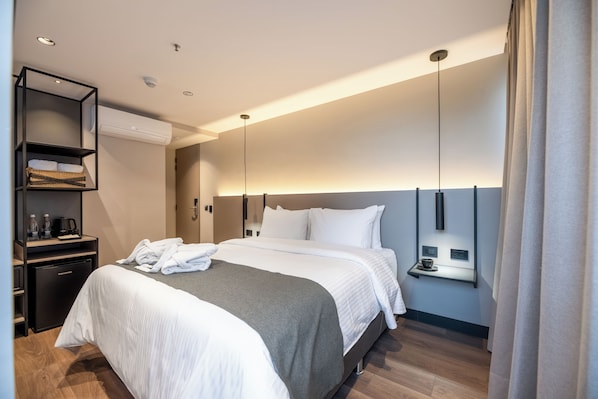 A modern hotel room with a neatly made bed, a minibar, a wall-mounted heater, and a bedside table with a cup and saucer. - BLOSS HOTEL (Medellín)