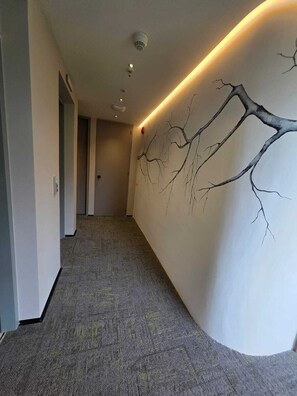 A hallway with a textured carpet, a wall-mounted tree branch artwork, recessed lighting, and a door at the end. - BLOSS HOTEL (Medellín)